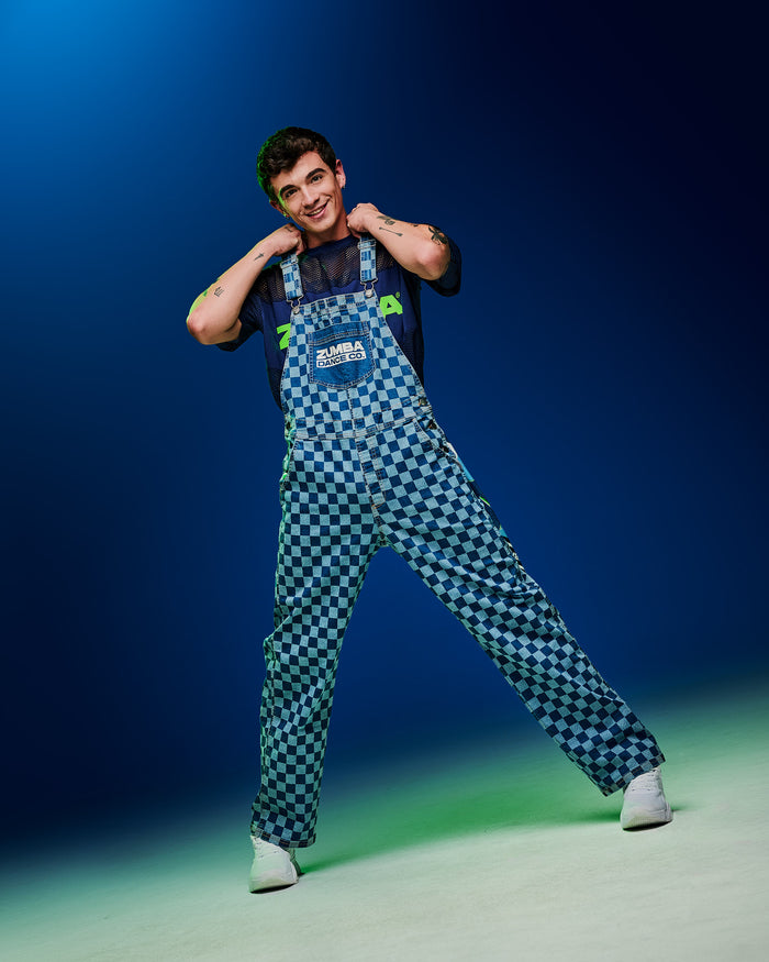 Male model posing with hands on his shoulders and feet apart, wearing checkered overalls against a blue-green gradient background.