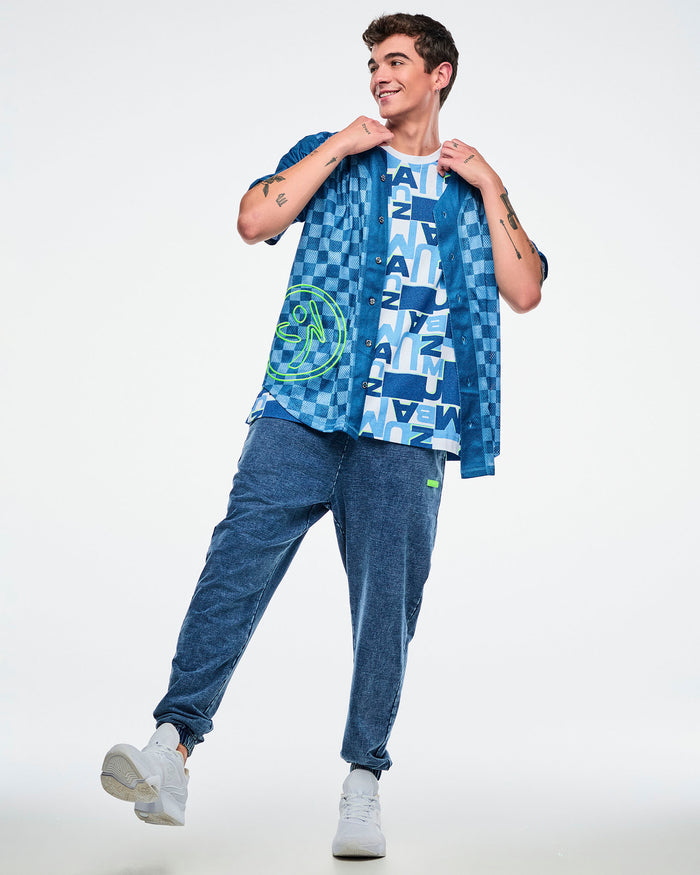 Full view of a male model wearing a blue patterned top and denim pants and white shoes on a white background.
