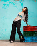 Black wide-leg Zumba pants with multicolor vertical “ZUMBA” logo on the lower leg; female model wears a white mesh Zumba top over a bra and white sneakers, leaning against stacked red, green, and blue crates in front of a turquoise wall.