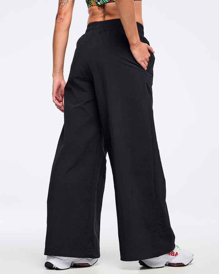 Back view of black wide-leg Zumba pants with elastic waistband and side pockets; female model wears white sneakers on a white background.