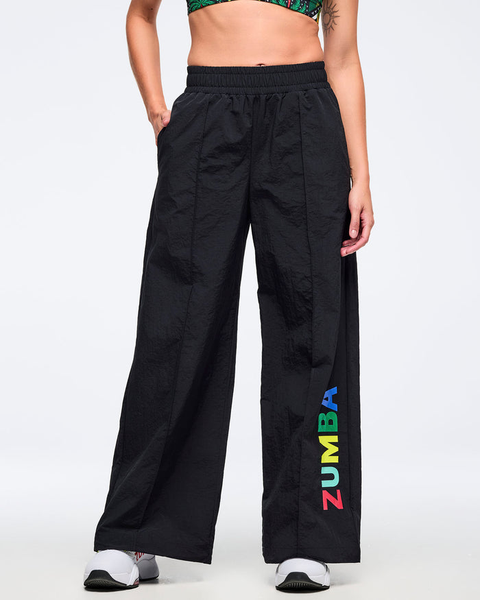 Front view of black wide-leg Zumba pants with side pockets and multicolor vertical “ZUMBA” logo near the hem; female model wears a tropical-print bra and white sneakers on a white background.