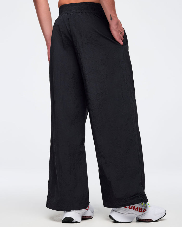 Back view of black wide-leg Zumba pants with elastic waistband and side pockets; male model wears white sneakers on a white background.