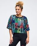 Multicolor Zumba mesh crop top with oversized fit, short sleeves, and black ribbed crew neck, covered in abstract “ZUMBA” lettering. Female model pairs it with high-waisted black leggings on a light gray studio background.