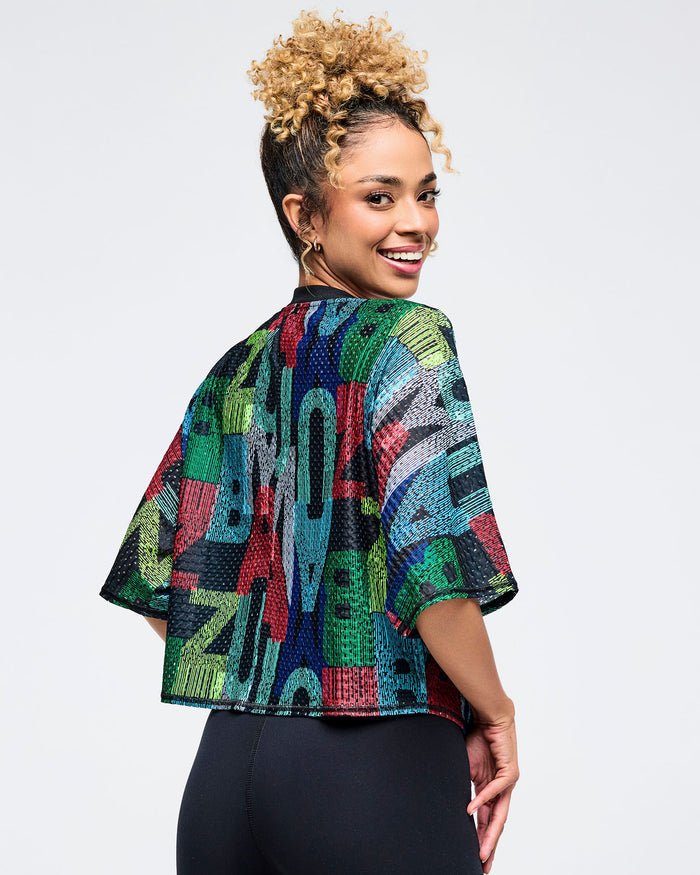Back view of the multicolor Zumba mesh crop top showing the allover abstract “ZUMBA” lettering print and boxy cropped hem. Female model wears it with high-waisted black leggings on a light gray studio background.