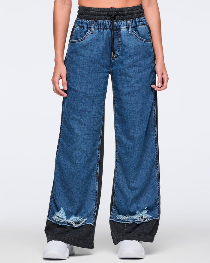Front view of wide-leg two-tone denim jogger pants with a blue denim front, black waistband and side panels, drawstring detail, front pockets, and distressed frayed hems, worn with white sneakers.