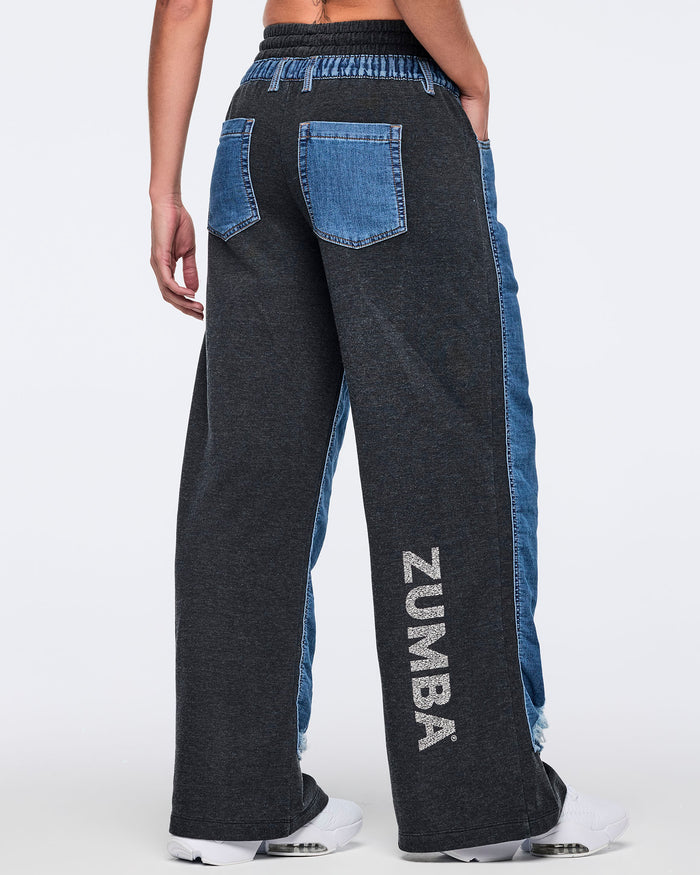 Back view of two-tone wide-leg denim jogger pants with a black body, blue denim side panels, elastic waistband with drawstring, denim back pockets, and vertical “ZUMBA” logo on the lower leg, worn with white sneakers.