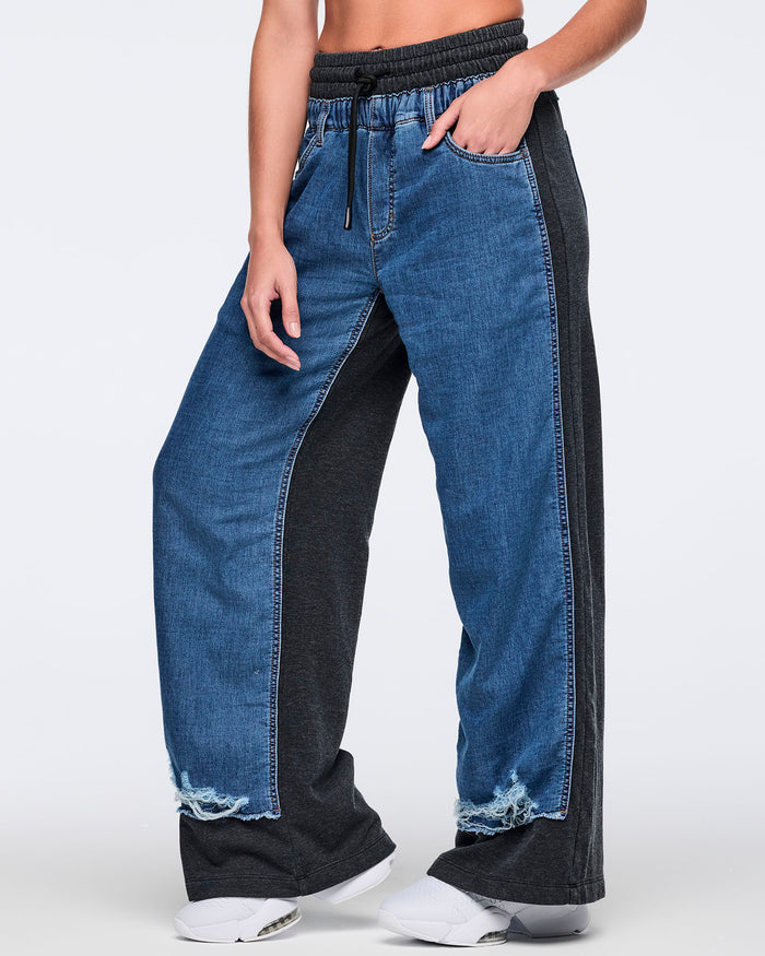 Side view of wide-leg two-tone denim jogger pants featuring blue denim front panels, black side/back panels, elastic drawstring waistband, front pockets, and frayed hems, paired with white sneakers.