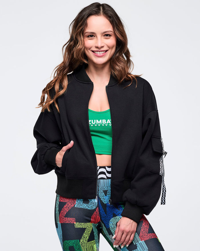 Black zip-up jacket worn open over a green “ZUMBA” scoop neck tank, styled with multicolor letter-print leggings; female model poses on a white background.