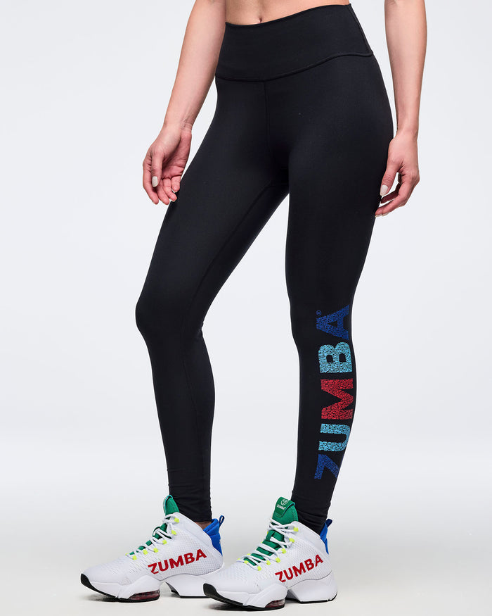High-waisted black leggings with vertical multicolor “ZUMBA” lettering down the lower leg; female model wears white high-top sneakers with white laces, shown on a white background.