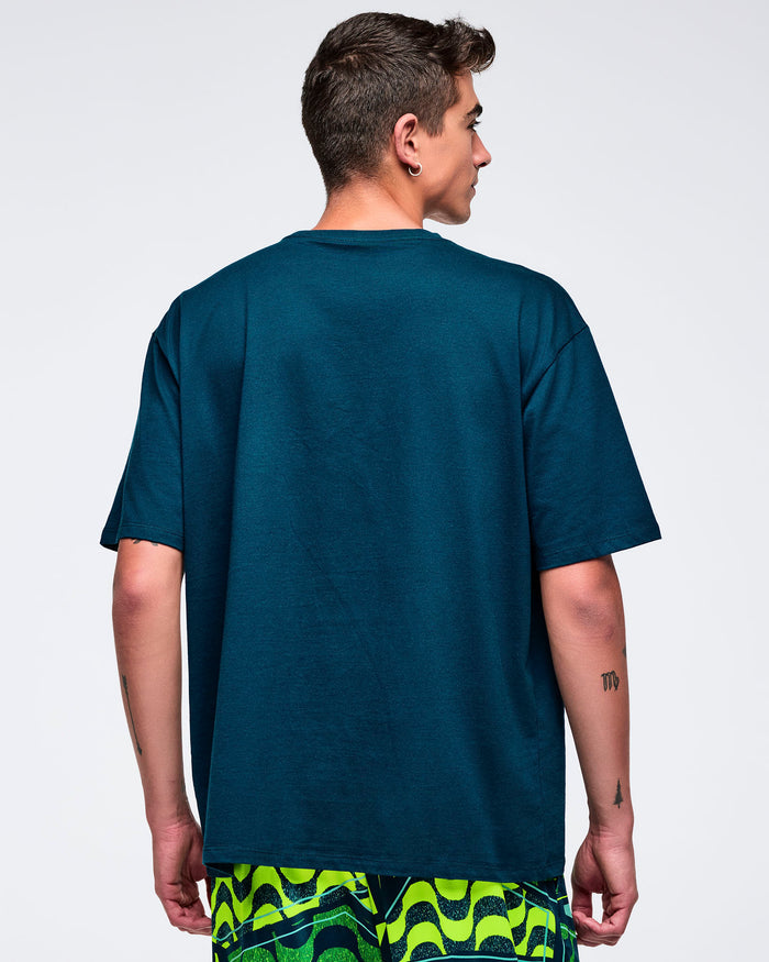 Back view of the dark teal oversized T-shirt on a male model, showing the plain back and relaxed drop-shoulder fit, paired with green patterned shorts.