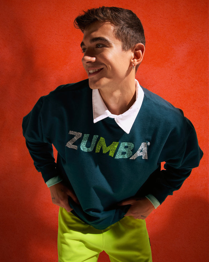 Teal crewneck sweatshirt with a distressed “ZUMBA” chest logo, modeled by a male wearing neon lime shorts over a white collared shirt against a textured orange backdrop.
