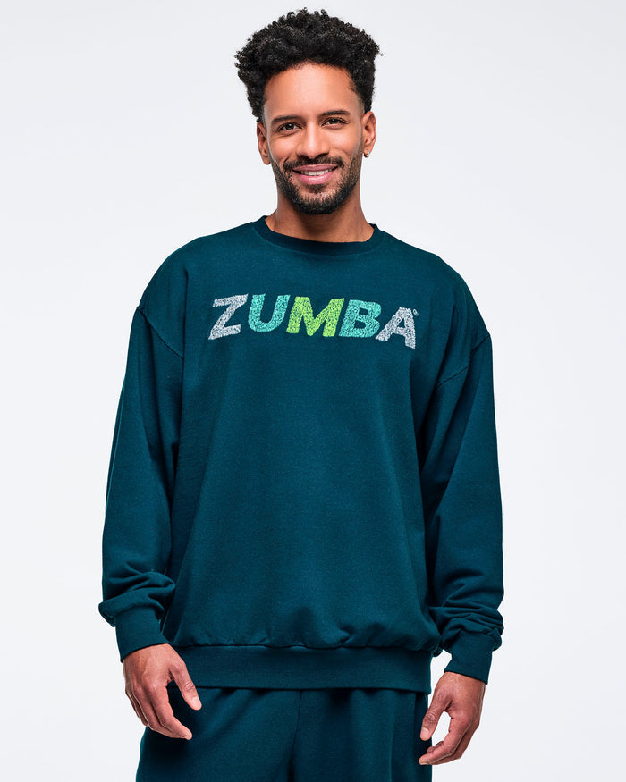 Teal oversized crewneck sweatshirt with ribbed cuffs and hem and a distressed “ZUMBA” logo on the chest, modeled by a male on a white studio background.