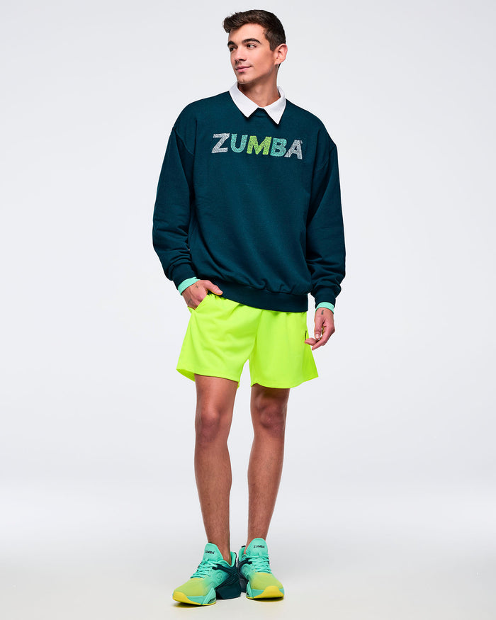 Teal oversized crewneck sweatshirt with a distressed “ZUMBA” logo across the chest and ribbed cuffs and hem, modeled by a male wearing a white collared shirt underneath, neon lime shorts, and teal-to-lime athletic sneakers on a white studio background.