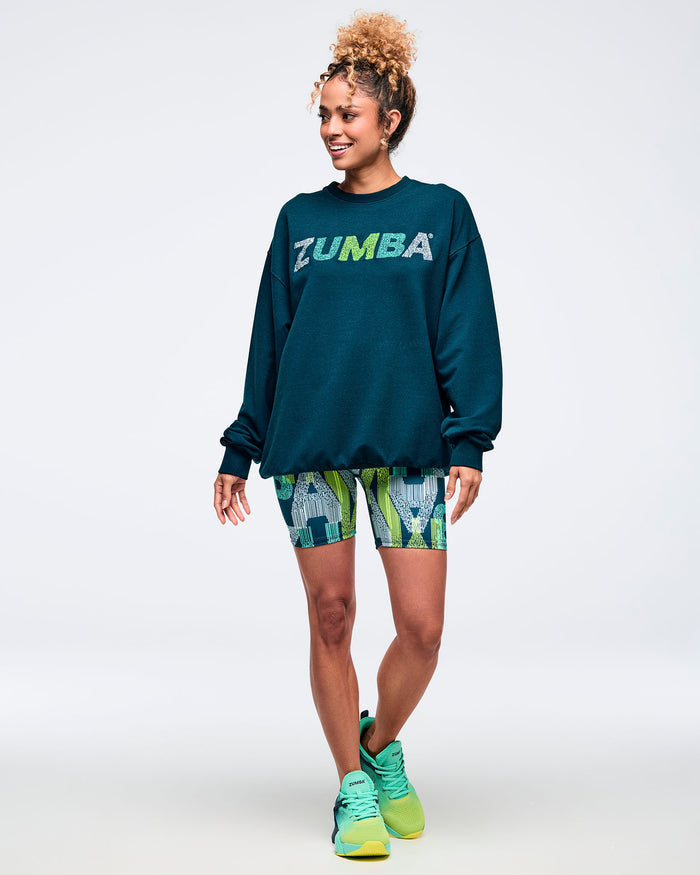 Female model wearing an oversized dark teal crewneck sweatshirt with multicolor “ZUMBA” logo across the chest, paired with fitted mid-thigh biker shorts in a teal, lime, and gray abstract print. She completes the look with teal-to-neon green lace-up athletic sneakers. Model stands smiling against a plain light background.