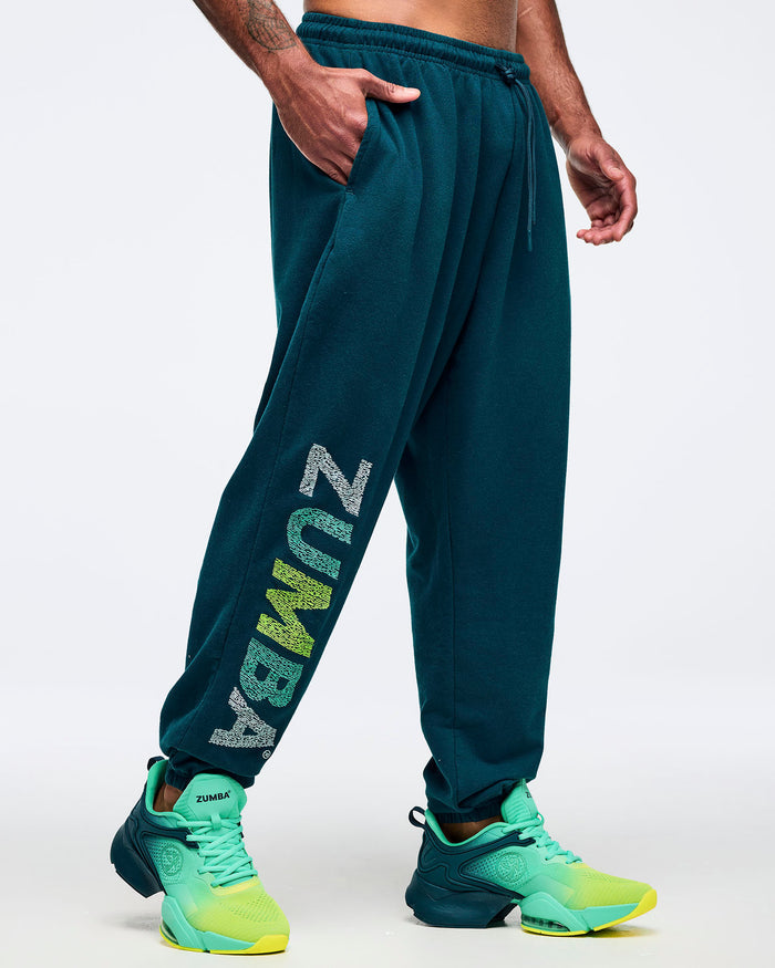 Teal jogger sweatpants with elastic drawstring waistband, side pockets, and a vertical multicolor “ZUMBA” graphic on the lower leg, modeled by a male wearing teal-to-lime athletic sneakers on a white studio background.