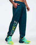 Teal jogger sweatpants with elastic drawstring waistband, side pockets, and a vertical multicolor “ZUMBA” graphic on the lower leg, modeled by a male wearing teal-to-lime athletic sneakers on a white studio background.