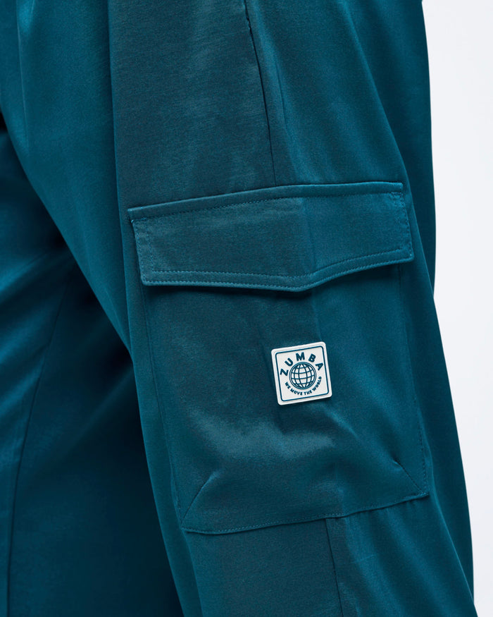 Close-up of teal cargo jogger pants showing the flap cargo pocket and stitched Zumba logo patch on the thigh.
