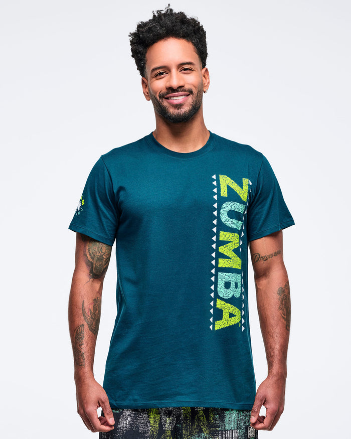 Teal short-sleeve crewneck T-shirt with a vertical lime “ZUMBA” graphic and white triangle border on the front, modeled by a male wearing patterned shorts on a white studio background.