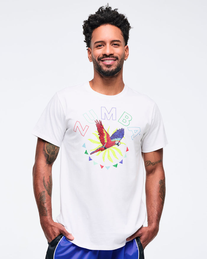 White short-sleeve graphic T-shirt with a colorful parrot and sunburst print and “ZUMBA” lettering across the chest, worn by a male model with bright blue track pants featuring black side panels and white piping.