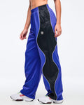 Blue and black track pants with white piping, elastic waistband, and wide straight-leg fit, shown from the side on a female model wearing white athletic sneakers.