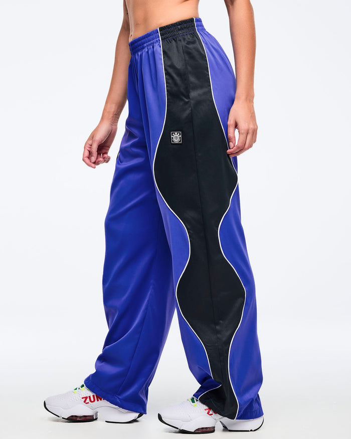 Blue and black track pants with white piping, elastic waistband, and wide straight-leg fit, shown from the side on a female model wearing white athletic sneakers.