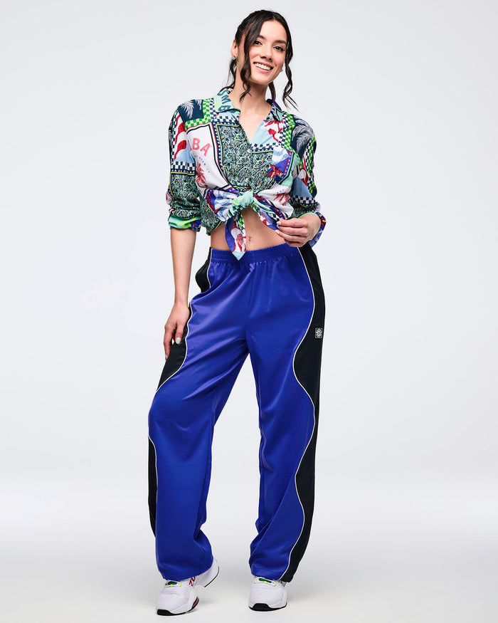 Blue track pants with black side panels, white piping, elastic waistband, and relaxed straight-leg fit, modeled by a female wearing a multicolor patchwork button-up shirt tied at the waist and white athletic sneakers on a white studio background.