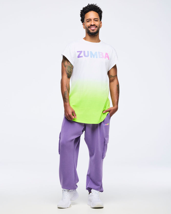 Full-body view of a man in a pastel green-to-white ombré “Zumba” T-shirt, pastel purple slouch cargo pants, and white shoes on a white background.