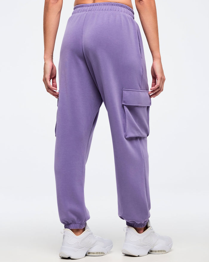 Back view of a female model wearing purple slouch cargo jogger pants with an elastic waistband, tapered elastic cuffs, and a side flap cargo pocket, paired with white sneakers on a white background.