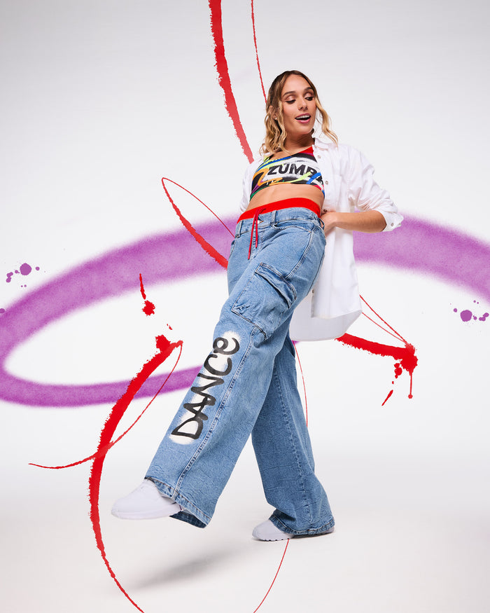 Female model in crop top and oversized white button down shirt with wide‑leg denim cargo pants that say “DANCE,” posing against a white backdrop with red and purple paint swirls.