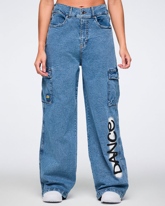 Female model wearing denim cargo pants with “DANCE” spray‑painted on a white stripe, standing against a white background.