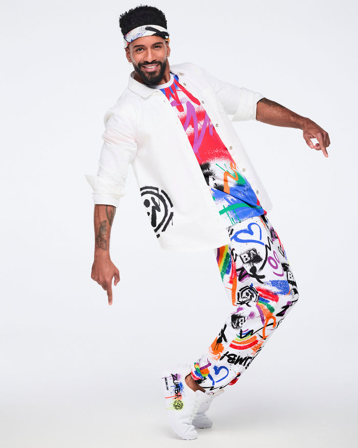 Male model wearing a bandana on this head, a white button‑down shirt, colorful graphic tee, matching graphic pants, and white Zumba shoes against a white background.