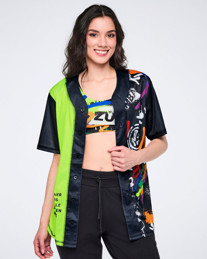 Female model in a neon green and black button-up Zumba jersey over a colorful sports bra and paired with black pants against a white background.