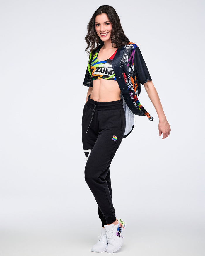 Female model in colorful Zumba crop top, black joggers, and white sneakers posing against a white background.