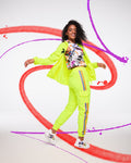 A female model dances while wearing a bright yellow jacket, a white graffiti-style hoodie, bright yellow pants with a rainbow stripe, and matching graffiti-style white shoes, surrounded by colorful paint-like designs on a white background.