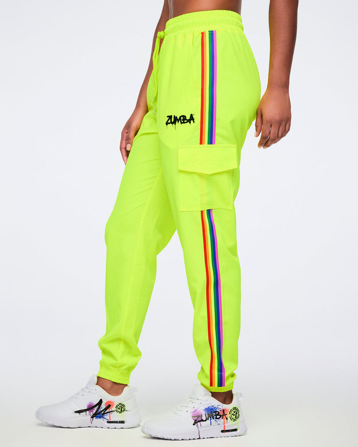 A female model wearing bright yellow pants featuring a graffiti-style “Zumba” logo on the upper left leg, a rainbow stripe down the side, and a cargo pocket, paired with white graffiti-style sneakers, posed against a plain white background.