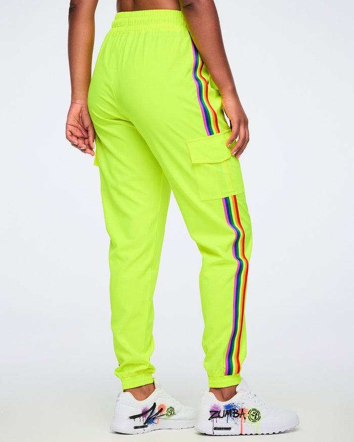 A female model wearing bright yellow pants with a rainbow stripe running down the leg and a side cargo pocket, paired with white graffiti-style sneakers, shown from the back against a plain white background.