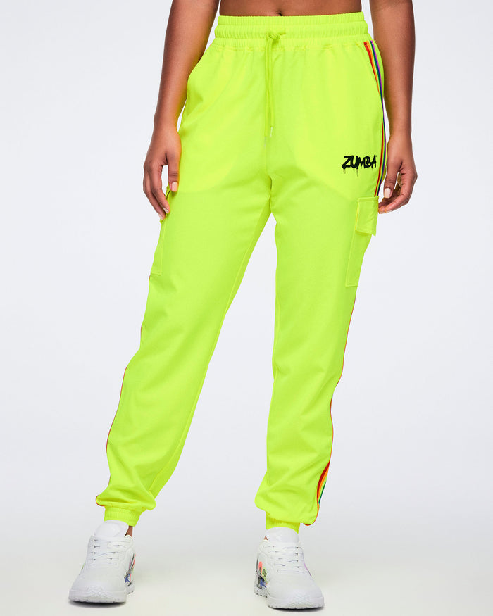 A female model wearing bright yellow pants with a graffiti-style “Zumba” logo on the upper left leg, a rainbow stripe down the side, and a cargo pocket, paired with white graffiti-style sneakers, against a plain white background.