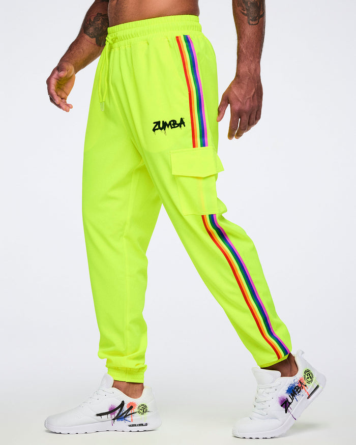 A male model wearing bright yellow pants featuring a graffiti-style “Zumba” logo on the upper left leg, a rainbow stripe down the side, and a cargo pocket, paired with white graffiti-style sneakers, against a plain white background.