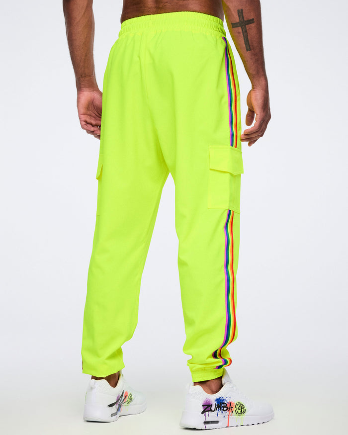 A male model shown from the back, wearing bright yellow pants featuring a rainbow stripe down the side, and a cargo pocket, paired with white graffiti-style sneakers, against a plain white background.