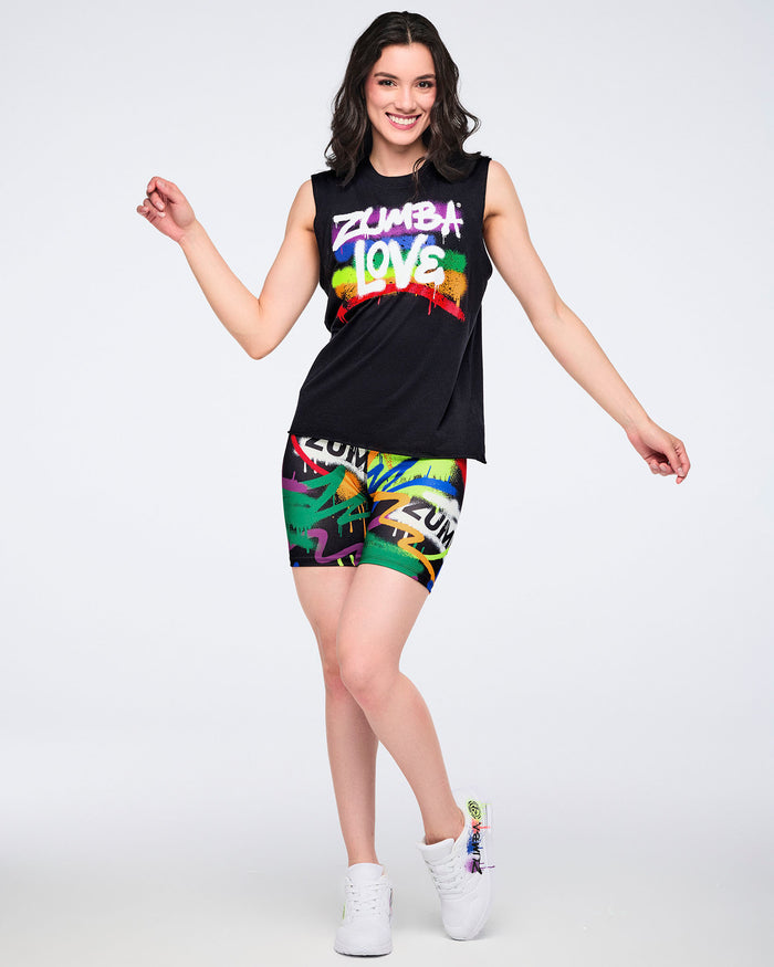 Full body view of a female model wearing a black sleeveless top with 'Zumba Love' text in white over a rainbow, paired with colorful biker shorts and white graffiti-style shoes on a white background.