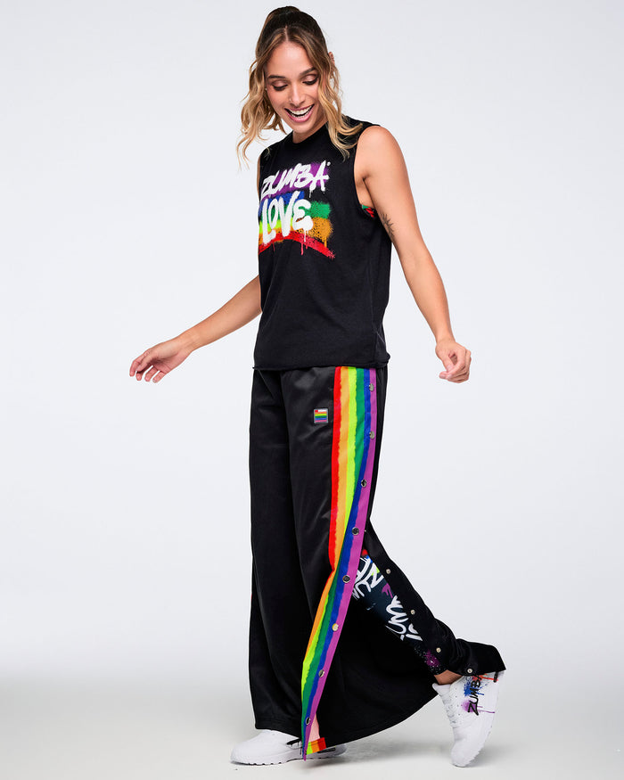Female model in a black sleeveless top with white “Zumba Love” over a rainbow, paired with black pants with rainbow stripes, unbuttoned to reveal colorful leggings and white graffiti-style shoes on a white background.