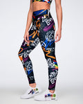 Female model wearing vibrant, graffiti‑inspired leggings featuring multicolored artwork on a black base, set against a white background.