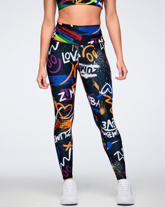 Female model wearing black high‑waisted graffiti‑print Zumba leggings with colorful neon artwork and white sneakers against a white background.