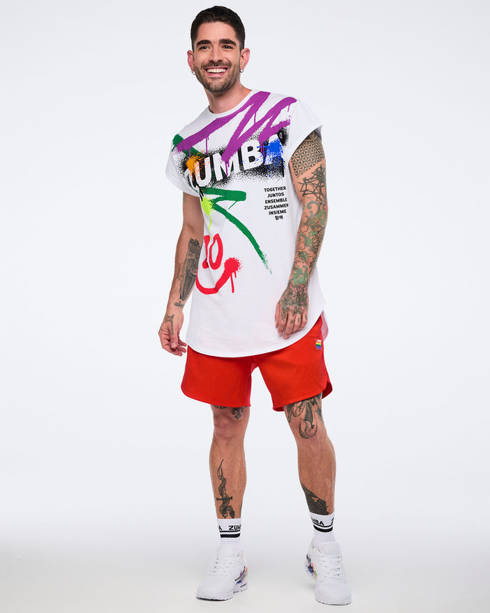 Male model wearing a colorful graphic t-shirt and red shorts and white 'Zumba' sneakers on a white background.