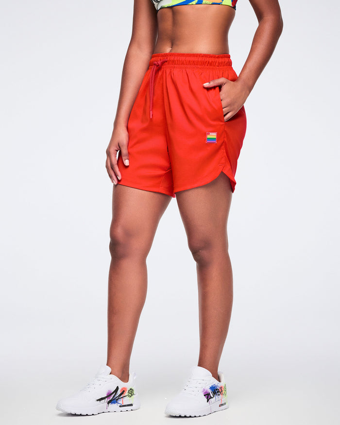 Female model wearing red shorts with a colorful logo and white 'Zumba' sneakers on a white background.