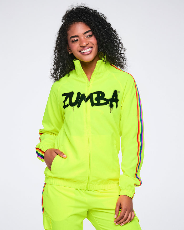 Female model wearing a bright yellow Zumba tracksuit with rainbow stripes on a sleeves, matching pants and a white background.
