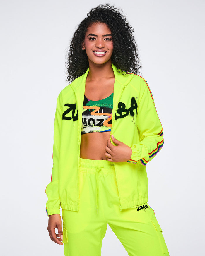 Female model wearing an open bright yellow jacket over a colorful Zumba‑branded bra, paired with matching bright yellow track pants on a white background.