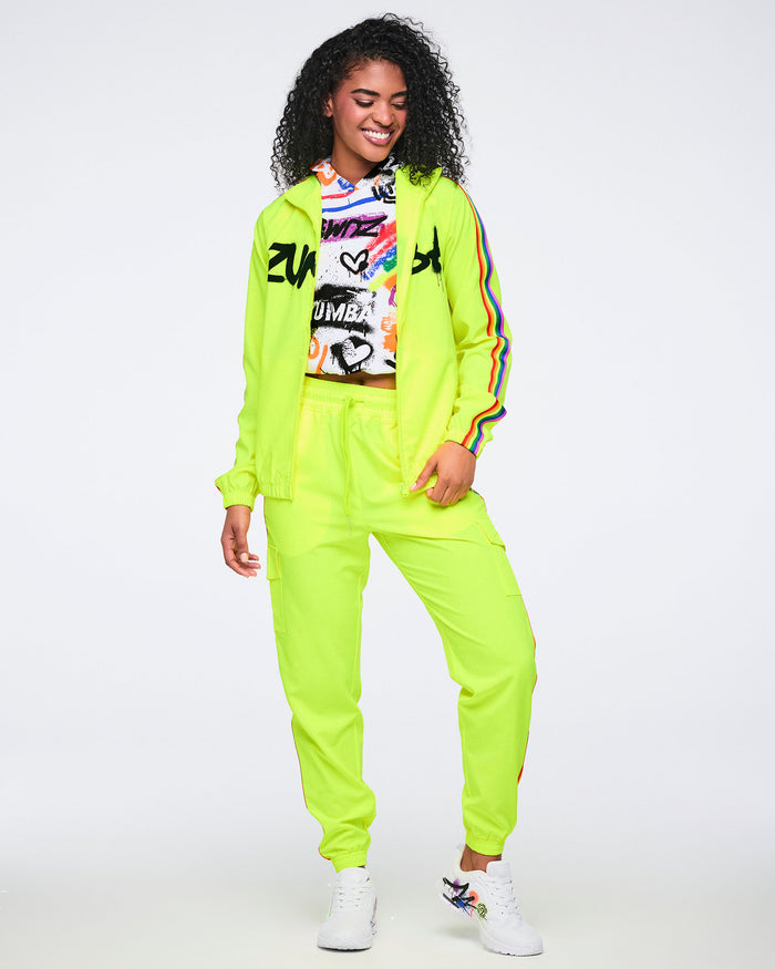 Female model wearing a bright yellow jacket with rainbow sleeve stripes over a colorful abstract hoodie, paired with bright yellow track pants featuring rainbow accents on a white background.