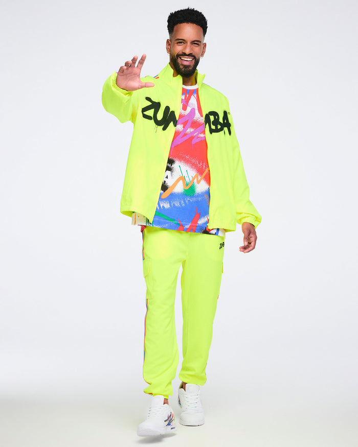 Male model wearing a bright yellow jacket over an abstract graphic T‑shirt, paired with bright yellow pants and white Zumba sneakers on a white background.