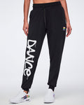 Female model wearing black joggers with bold white “DANCE” lettering down her right leg and white graffiti‑accented sneakers against a white background.